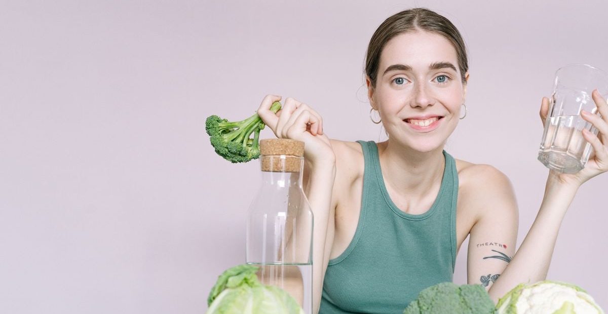 Woman in tank top smiling with broccoli, cabbage, and glass of water on table.