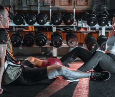 A man and woman resting together on the gym floor, surrounded by dumbbells.